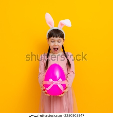 Cute baby little girl with bunny ears holding a big Easter egg on a colored yellow background.
