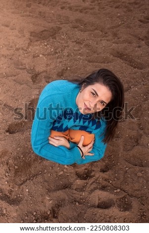 Girl posing on sand in the garden, dressed in saturated colors, blue sweater and orange pants, shot against a garden background.