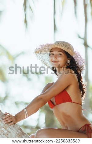 Summer lifestyle funny portrait of cute young bikini girl with tanned perfect body in red swimsuit, posing sitting on palm tree and looking at the camera at the tropical beach resort during sea