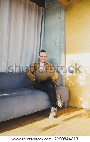 Young male worker in casual clothes and glasses looking at laptop while sitting on sofa with laptop in bright modern workspace. A successful freelancer works remotely online in a workspace
