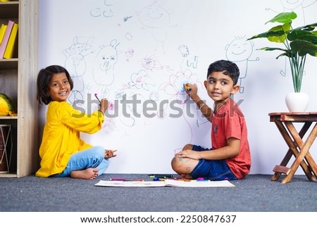 Young siblings kids drawing with crayon color on wall by looking at camera at home - concept of mischief, troublesome children and childhood lifestyles.