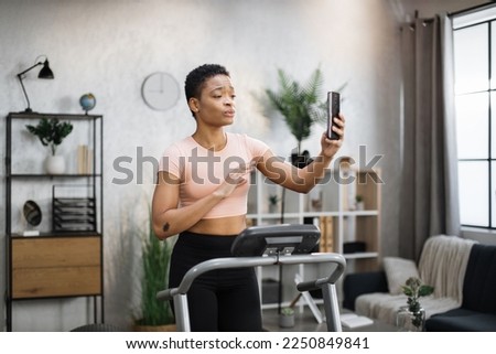 Attractive young sports african woman having online video call while working out, running, doing cardio training on treadmill in morning time, indoor on background of modern apartment or gym.