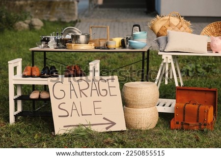 Sign Garage sale written on cardboard near tables with different stuff in yard