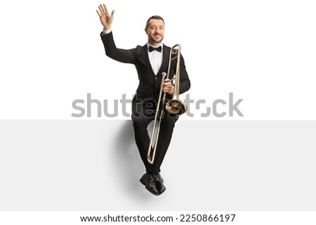 Male musician sitting on a blank panel with a trombone and waving isolated on white background Royalty-Free Stock Photo #2250866197