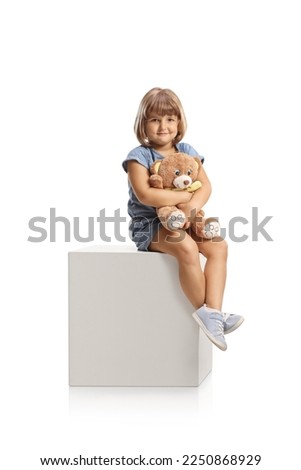 Cute little girl holding a teddy bear and sitting on a white cube isolated on white background Royalty-Free Stock Photo #2250868929
