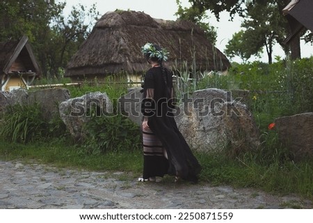 Ukrainian aesthetic scenic photography. Picture of woman in black folk dress wearing leaves wreath with hut on background. High quality wallpaper. Photo concept for ads, travel blog, magazine, article