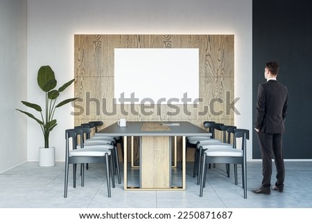Businessman back view looking at blank white poster on wooden wall decoration background in sunlit spacious meeting room with dark conference table on concrete floor, mockup