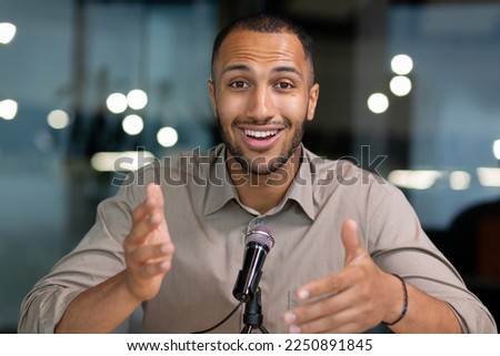 Successful hispanic man inside office looking at camera and talking on video twin inside office, businessman recording audio podcast using professional microphone, webcam view pov.