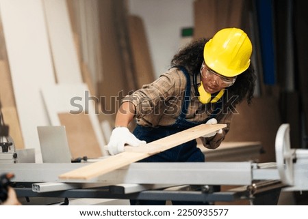Young attractive black female carpenter wearing safety goggles and hard hat for woodworking in small business workshop. Wood  industry and furniture industry.