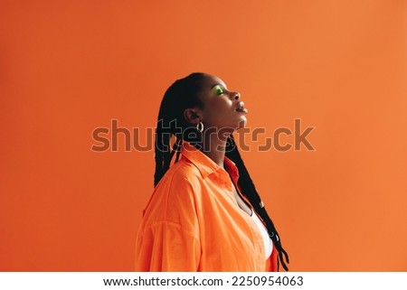 Fashionable african woman with dreadlocks standing in a studio with her eyes closed. Confident black woman wearing piercing jewellery and makeup. Female hipster standing against an orange background. Royalty-Free Stock Photo #2250954063