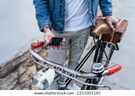 Cropped image of a young man in denim jacket holding his vintage classic bicycle while looking his mobile phone in front of a brick wall.