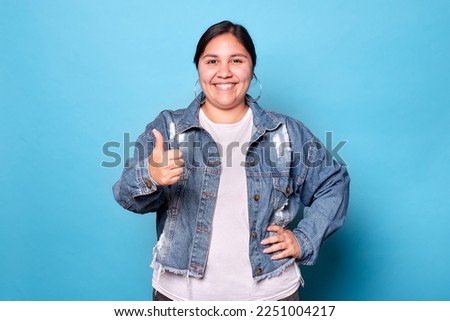 Young curvy latina woman wearing denim jacket and hoop earrings, smiling showing thumb up looking at camera isolated on blue background. Copy space.