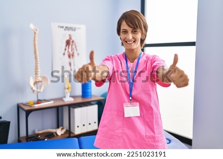 Brunette woman working at rehabilitation clinic approving doing positive gesture with hand, thumbs up smiling and happy for success. winner gesture. 