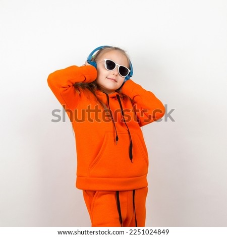 A little girl in an orange suit, sunglasses and blue headphones listens to music. Portrait on white background.