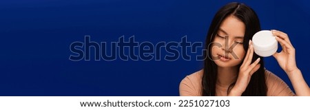 Brunette asian woman holding cosmetic cream isolated on blue, banner