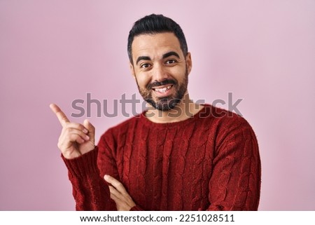 Young hispanic man with beard wearing casual sweater over pink background with a big smile on face, pointing with hand and finger to the side looking at the camera. 