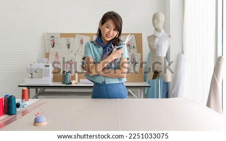 Young dressmaker in striped scarf holding scissor with a smile. Pin cushion, silk thread and ruler are on the workbench. Asian woman working in home base cloth design business.