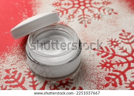 Winter skin care. Hand cream near snowflake silhouettes made with artificial snow on red background, closeup