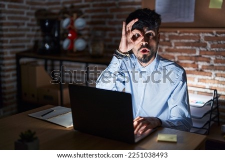 Young hispanic man with beard working at the office at night doing ok gesture shocked with surprised face, eye looking through fingers. unbelieving expression. 