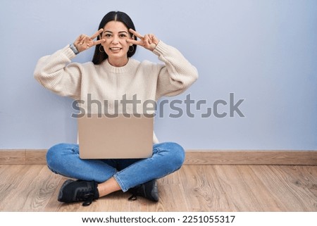 Young woman using laptop sitting on the floor at home doing peace symbol with fingers over face, smiling cheerful showing victory 