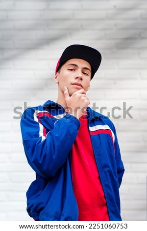 A teenage boy agains white brick wall. A male teenager model wearing casual clothing. A male teenager model wearing baseball uniform. Red t shirt and blue shirt. Street lifestyle clothing. Royalty-Free Stock Photo #2251060753