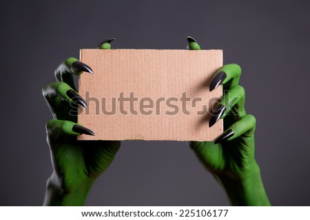 Green hands with black nails holding empty piece of cardboard, Halloween theme  