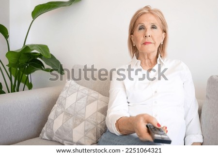 Cheerful senior woman enjoys evening with a TV shows, series. Smiling mature woman sits on the sofa with TV remote controller and switching channels
