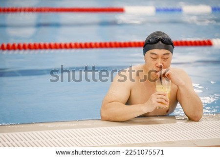 Asian man in hat and goggles drinking an orange juice in swimming pool