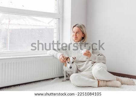 Relaxed, serene adult woman near radiator drinking cup of coffee in the living room. Middle-aged lady sitting with dog on floor at home  Royalty-Free Stock Photo #2251084105