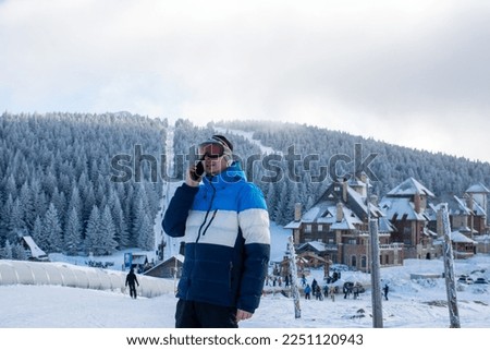 Happy man using smartphone in snowy mountains after skiing. Smiling guy talking with someone with copy space. Cheerful sporty man enjoy winter vacation at mountain with cellphone.