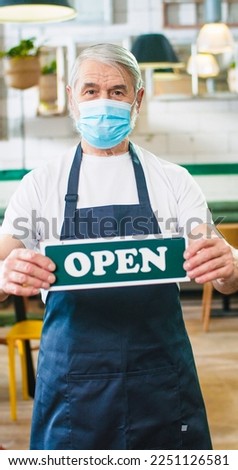 Portrait of old nice grey-haired Caucasian male worker in apron wearing medical mask looking at camera standing in coffee house holding in hands Open sign business in lockdown