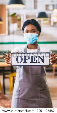 Portrait of pretty young African American young female employee in apron wearing medical mask standing in coffee shop holding in hands Open sign, reopening in quarantine, business