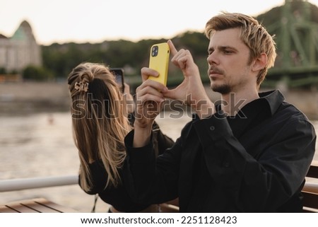 Woman and man enjoying beautiful landscape view on the riverside from the boat and takes picture using they mobile phones. Adventure, couple trip on boat and joy of city view.