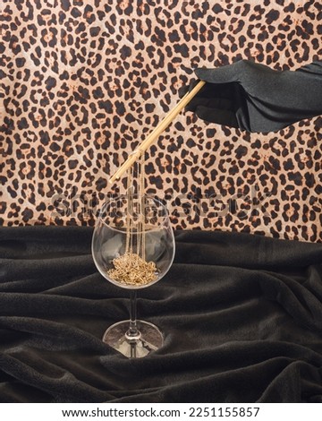 Black gloved hand holding chopsticks, picking gold necklace from wine glass against black and cheetah print background. Angled composition.