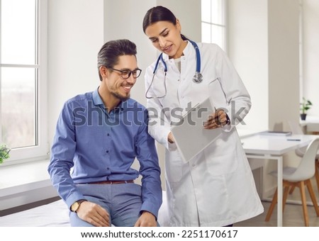 Professional female doctor smiles and shows positive examination results to young, joyful man. Handsome, happy guy with glasses at doctor's appointment in modern office of medical institution. Royalty-Free Stock Photo #2251170617