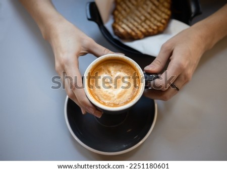 Thin female fingers hold a cup of coffee with heart shaped latte art foam. Close up cup of coffee with cream in coffee shop.
