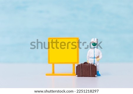 Miniature nurse in protective medical suit with luggage and blank notice board on blue background, scientist wearing protection suite to protect against coronavirus covid-19