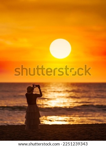 Young women come to travel to relax at the sea on vacation using A smartphone takes pictures of the sunset silhouette alone in the evening. At Khao Lak Beach, Phangnga, Thailand.