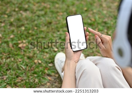 Young Asian woman in casual clothes wearing wireless headphones and using her mobile phone while relaxing on grass in the public park. phone white screen mockup. back view
