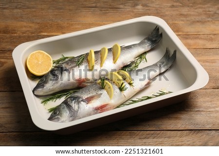 Baking tray with raw sea bass fish, lemon and rosemary on wooden table Royalty-Free Stock Photo #2251321601