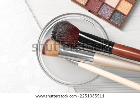 Cleaning makeup brushes in bowl with special liquid on white wooden table, flat lay Royalty-Free Stock Photo #2251335511