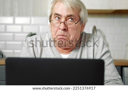 Senior grey haired man wearing glasses sitting at laptop and making fish face with lips. Crazy and comical gesture. Funny Old grandfather with playful silly facial expression grimacing fooling around