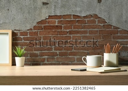 Stylish workplace with picture frame, cup of coffee, pencil holder and notebook on wooden table against brick wall
