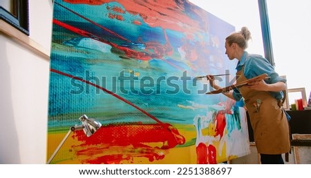 Side view of Caucasian young pretty talented female painter working in art studio drawing on big canvas. Modern creating painting on large canva. Woman professional artist in apron working in workshop