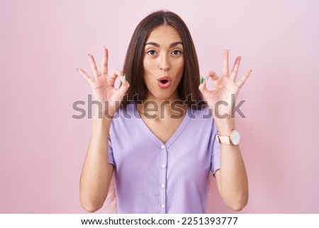 Young hispanic woman with long hair standing over pink background looking surprised and shocked doing ok approval symbol with fingers. crazy expression 