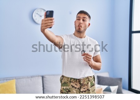 Young hispanic man wearing camouflage army uniform taking selfie at home making fish face with mouth and squinting eyes, crazy and comical. 