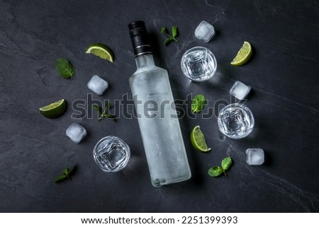 Bottle of vodka, shot glasses, lime, mint and ice on black table, flat lay Royalty-Free Stock Photo #2251399393
