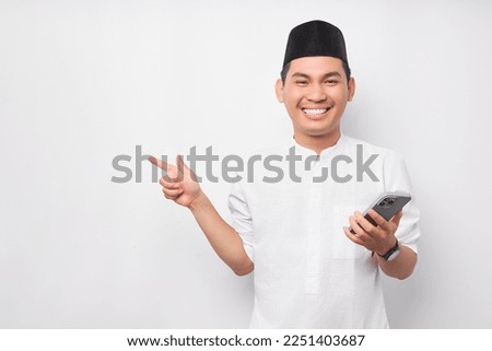 Smiling young Asian Muslim man holding mobile phone and pointing finger at empty copy space isolated on white background. People religious Islam lifestyle concept. celebration Ramadan and ied Mubarak