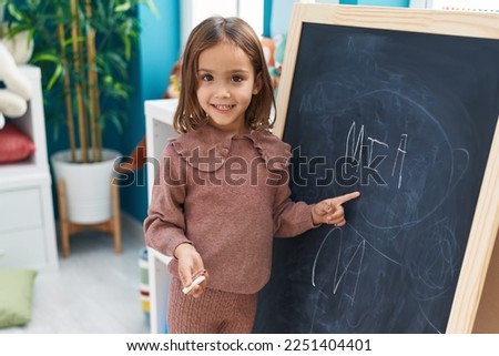 Adorable hispanic girl preschool student smiling confident writing name on blackboard at kindergarten Royalty-Free Stock Photo #2251404401