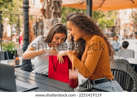 Two girls are sitting in the cafe, pulling out clothes from the shopping bag, and seem thrilled. Royalty-Free Stock Photo #2251419119